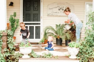 woman with young son and daughter on bungalow front porch watering potted plants