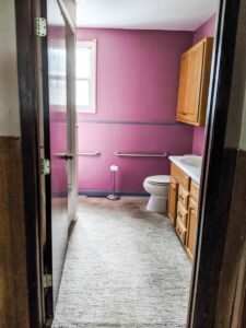 looking through doorway of farmhouse bathroom prior to remodel with purple wall and carpeted floors