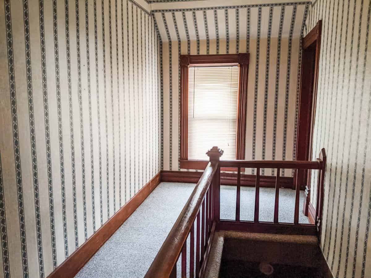 upstairs farmhouse hallway with wallpaper carpeted floor and original woodwork looking toward window