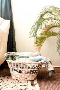 white vintage basket filled with quilts sitting on top of a rug next to a couch and potted plant