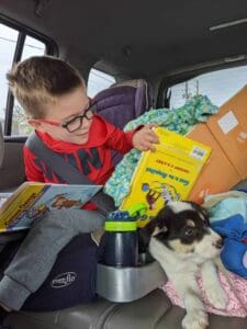 a little boy looking at books while strapped in his car seat with his puppy sitting next to him