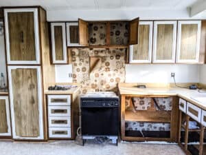 mobile home kitchen prior to renovation with peeling wallpaper partially painted cabinets and an uninstalled dishwasher