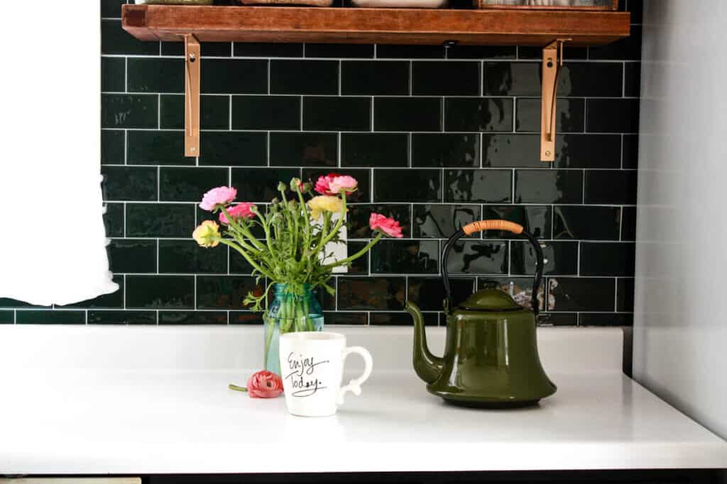 white kitchen countertop with green sticky tile open shelves fresh cut flowers coffee mug and a tea kettle
