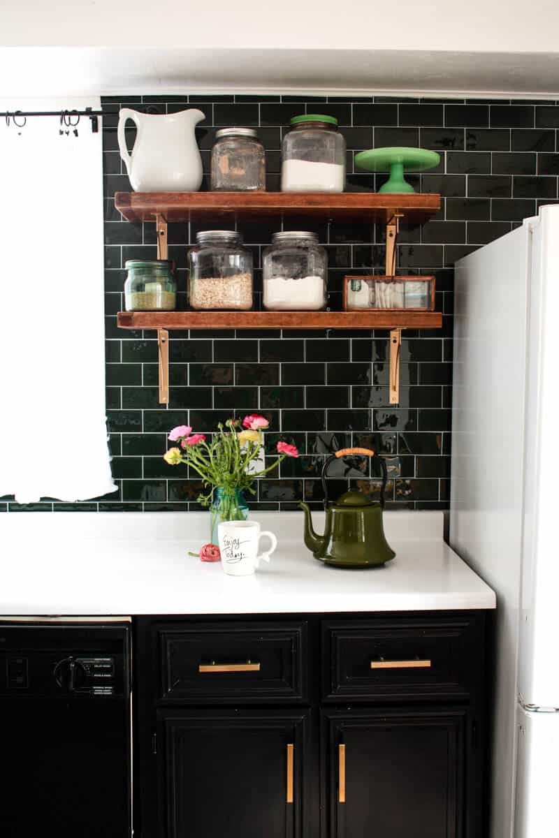 mid century modern kitchen with dark green sticky tile white countertops black cabinets open shelves and a white refrigerator