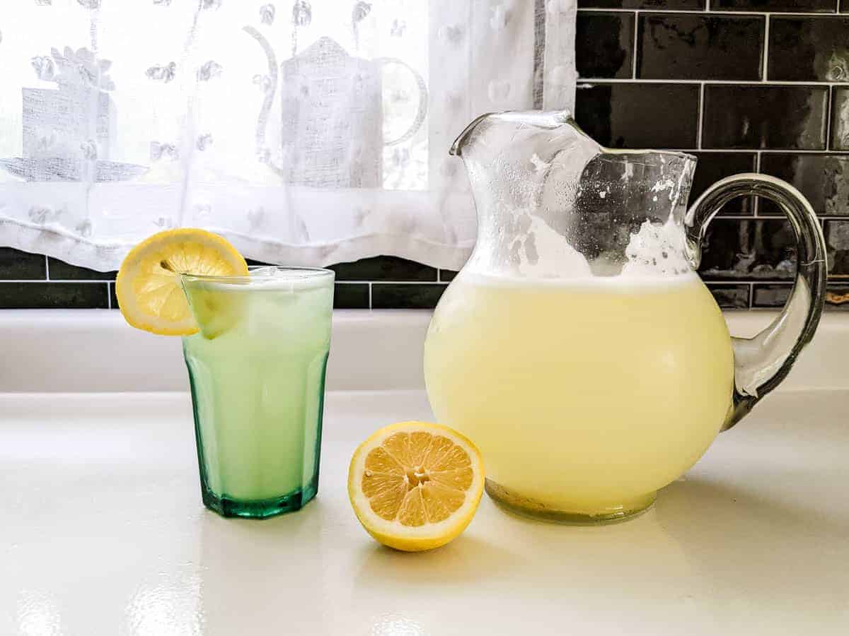 glass pitcher of homemade lemonade with half lemon and glass of lemonade sitting on kitchen counter