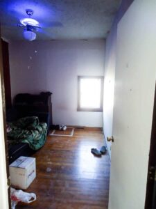 looking into dark bedroom before remodel with wooden floors white walls and small window