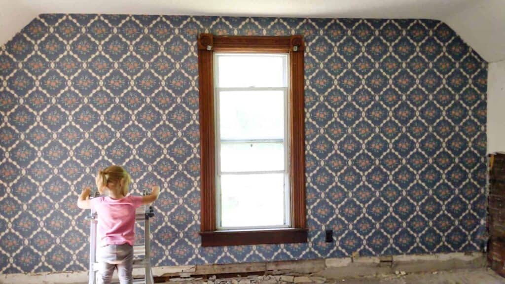 little girl standing in front of blue floral wallpapered farmhouse bedroom wall with window