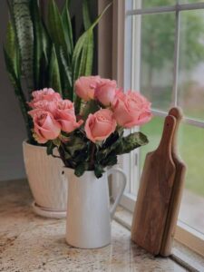 white pitcher full of pink roses on kitchen counter with wooden cutting board and potted snake plant