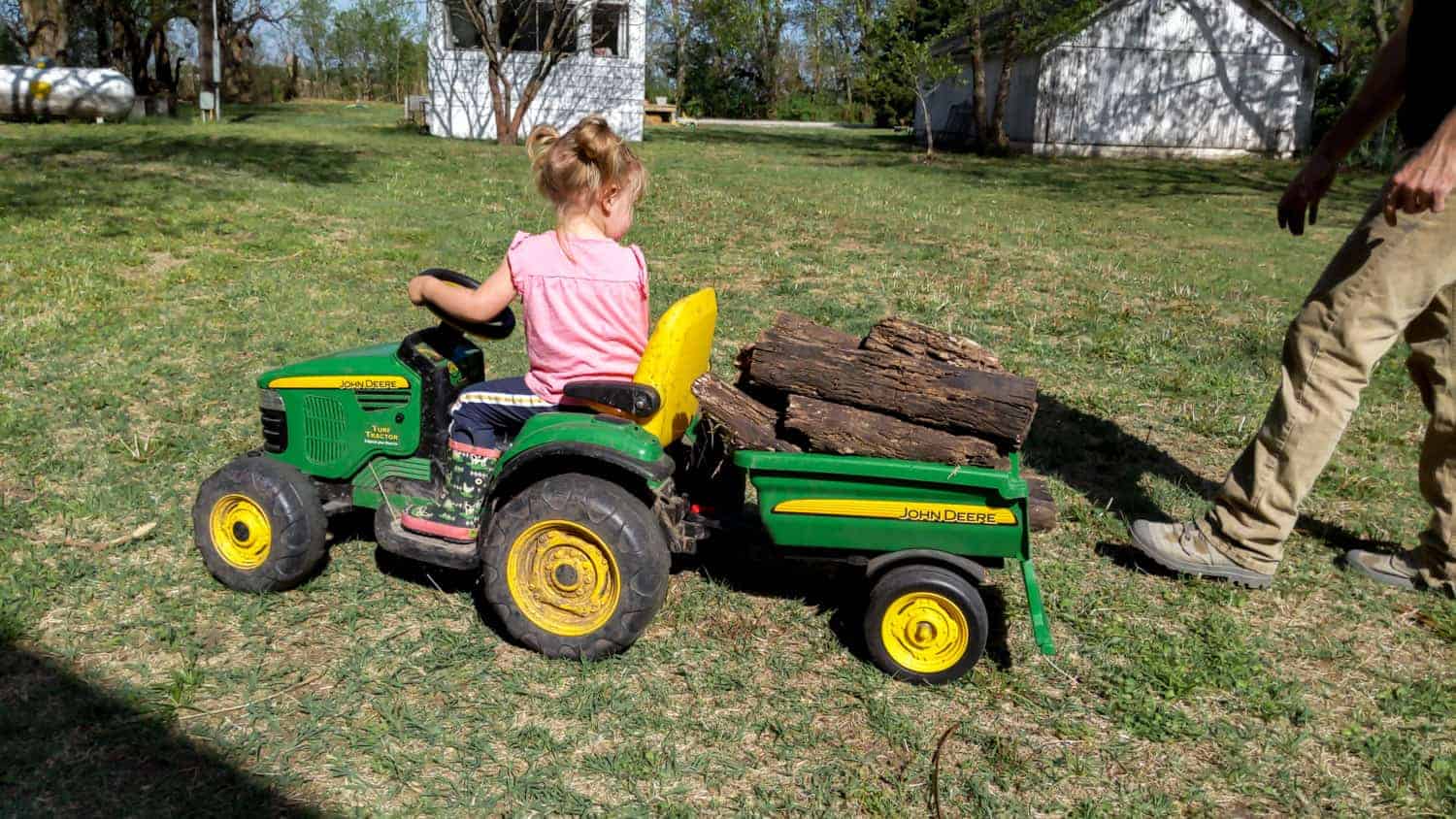 young girl drives a john deere ride on toy hauling fire wood around homestead